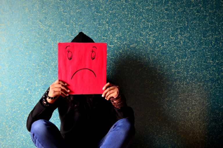Picture of a person wearing a black hoodie and jeans holding a red square board in front of their face. The board has a sad face drawn on it.